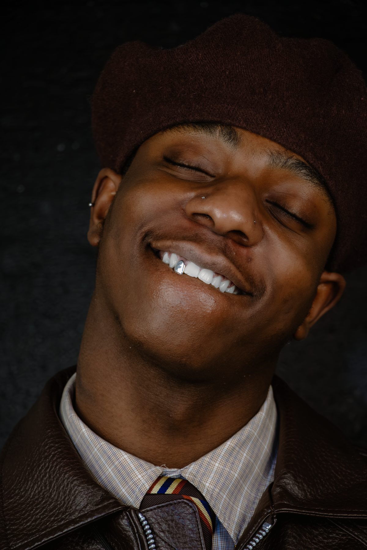 Shane in a brown beret, eyes closed, smiling to show a single silver cap